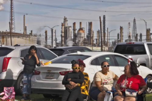 A group of women and children sit in chair and lean on a car in a parking lot. In the background, many smokestacks and tall exhaust vents send smoke into the air.