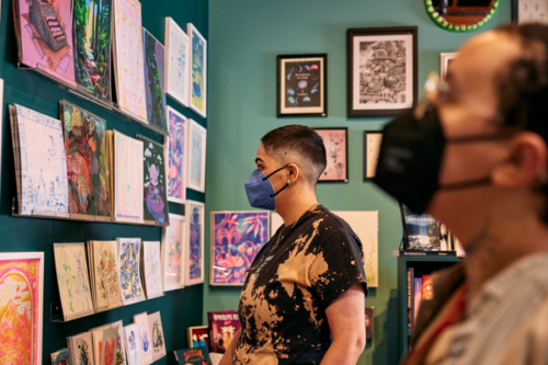 A masked person looks at a wall of artwork and books. Another masked person is in the foreground and out of focus.