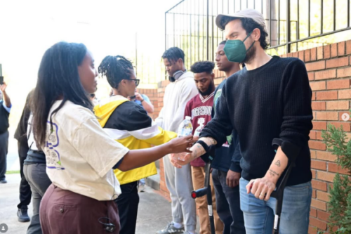 a black woman with dreads takes a bottle of water from a masked white man wearing a blue shirt, jeans, and a white hat.