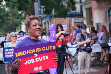 A black woman with short brown hair holds a sign that reads