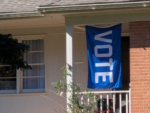 A large blue flag with white letters that read VOTE hangs on a porch of a brick and tan home.